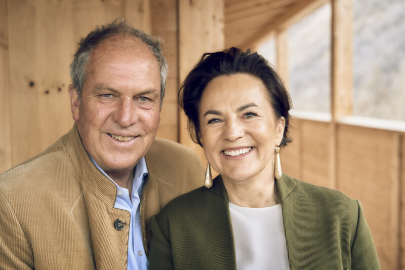 Portrait of a smiling man and woman, both dressed in elegant attire, against a wooden background.