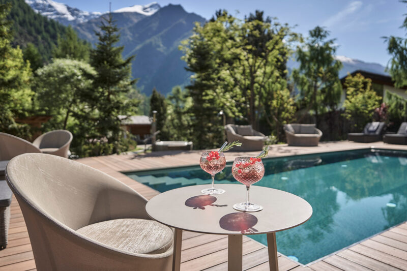 Two cocktails with garnishes on a table beside an outdoor pool, surrounded by trees and mountains in the background.