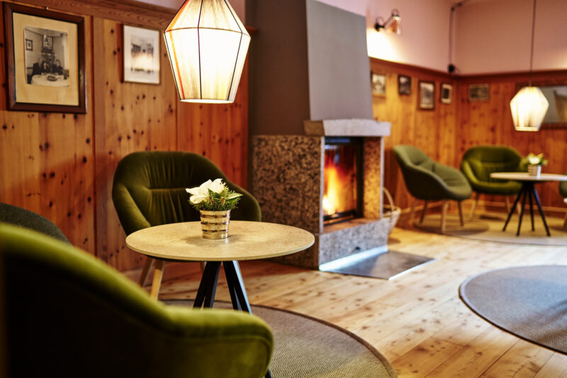 Cozy restaurant interior with green chairs, wooden walls, and a fireplace, featuring tables with floral centerpieces.