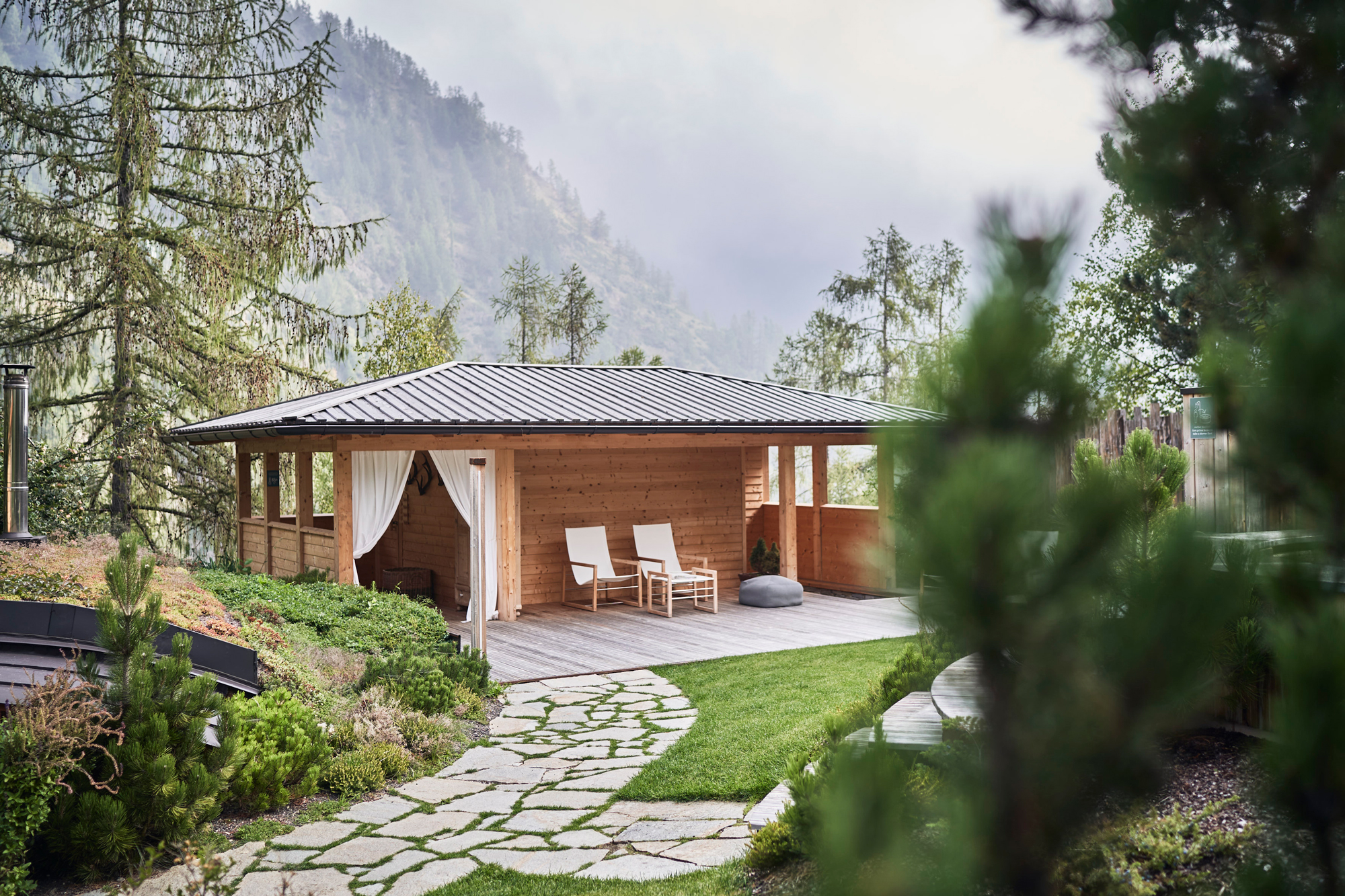 Wooden gazebo with two white chairs on a deck, surrounded by greenery and mountains in the background.