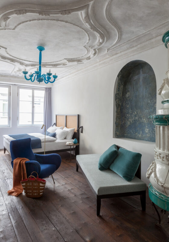 Elegant bedroom featuring a blue chandelier, a blue chair, a gray daybed with teal cushions, and a decorative wall niche.