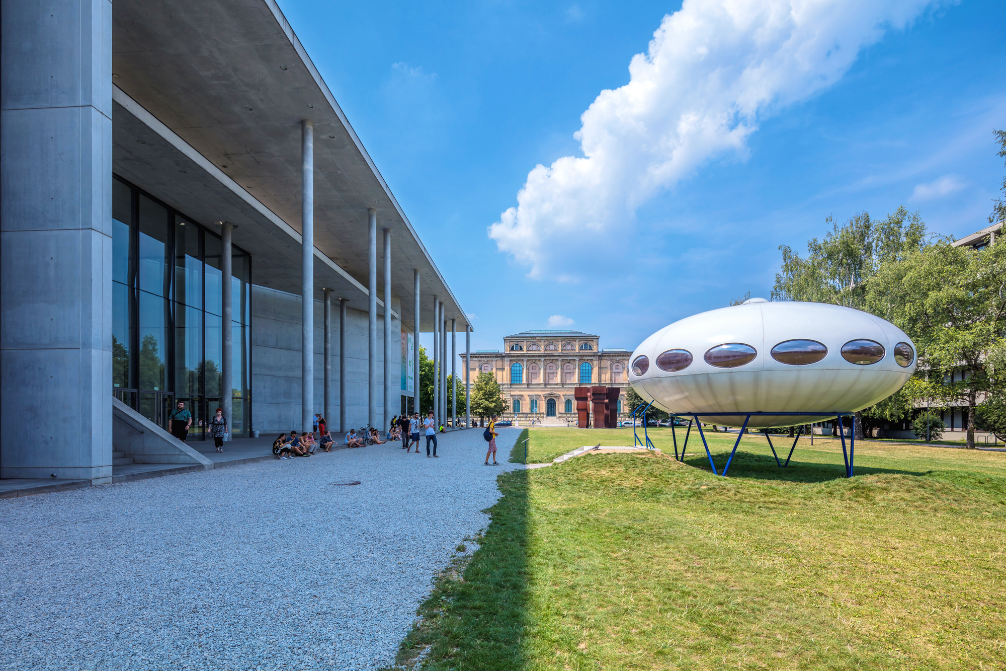 Modern building with large glass windows beside a futuristic, oval-shaped structure on a grassy area under a blue sky.