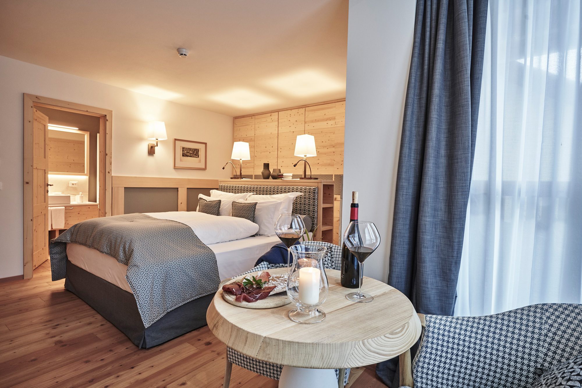Cozy hotel room featuring a bed, wooden accents, a small table with wine and snacks, and large windows with curtains.