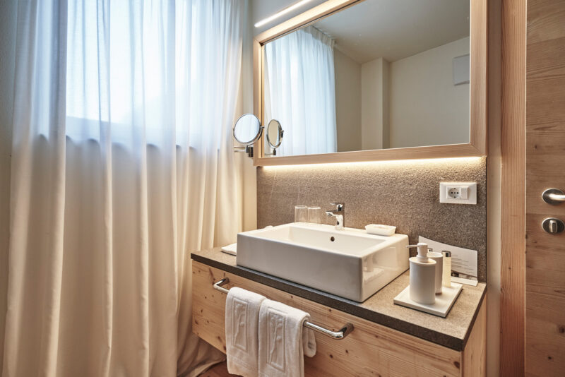 Modern bathroom with a white sink, wooden cabinetry, and large mirror, illuminated by natural light through sheer curtains.