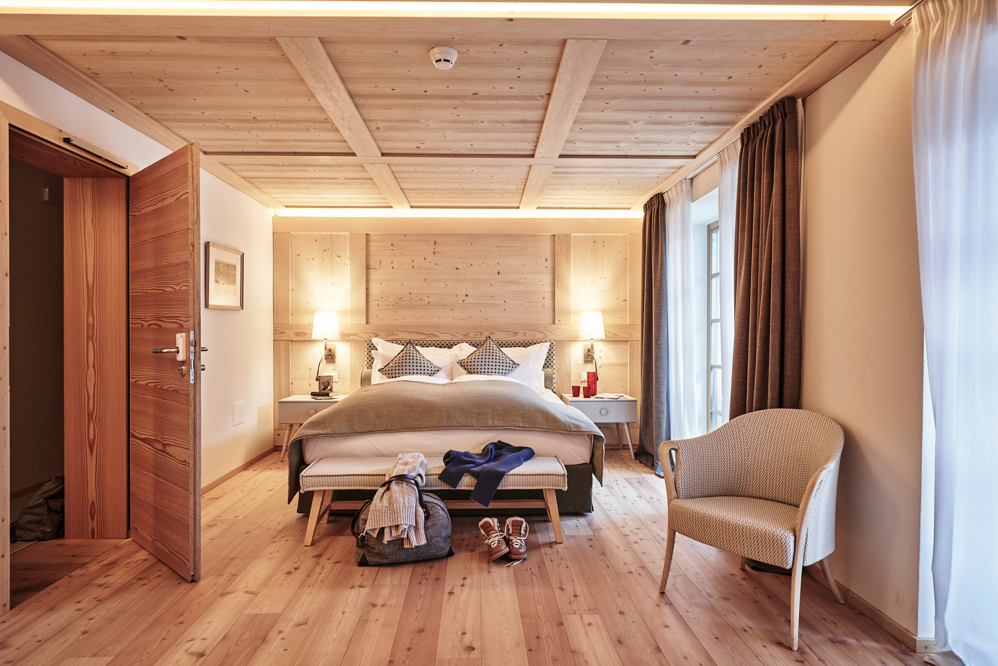 Deluxe room with wooden decor, a bed with decorative pillows, and a chair; luggage and clothing on the floor.