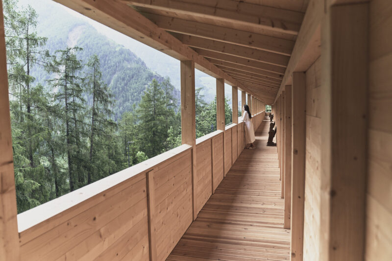 Wooden balcony overlooking a forested mountain landscape, with two people standing along the railing.