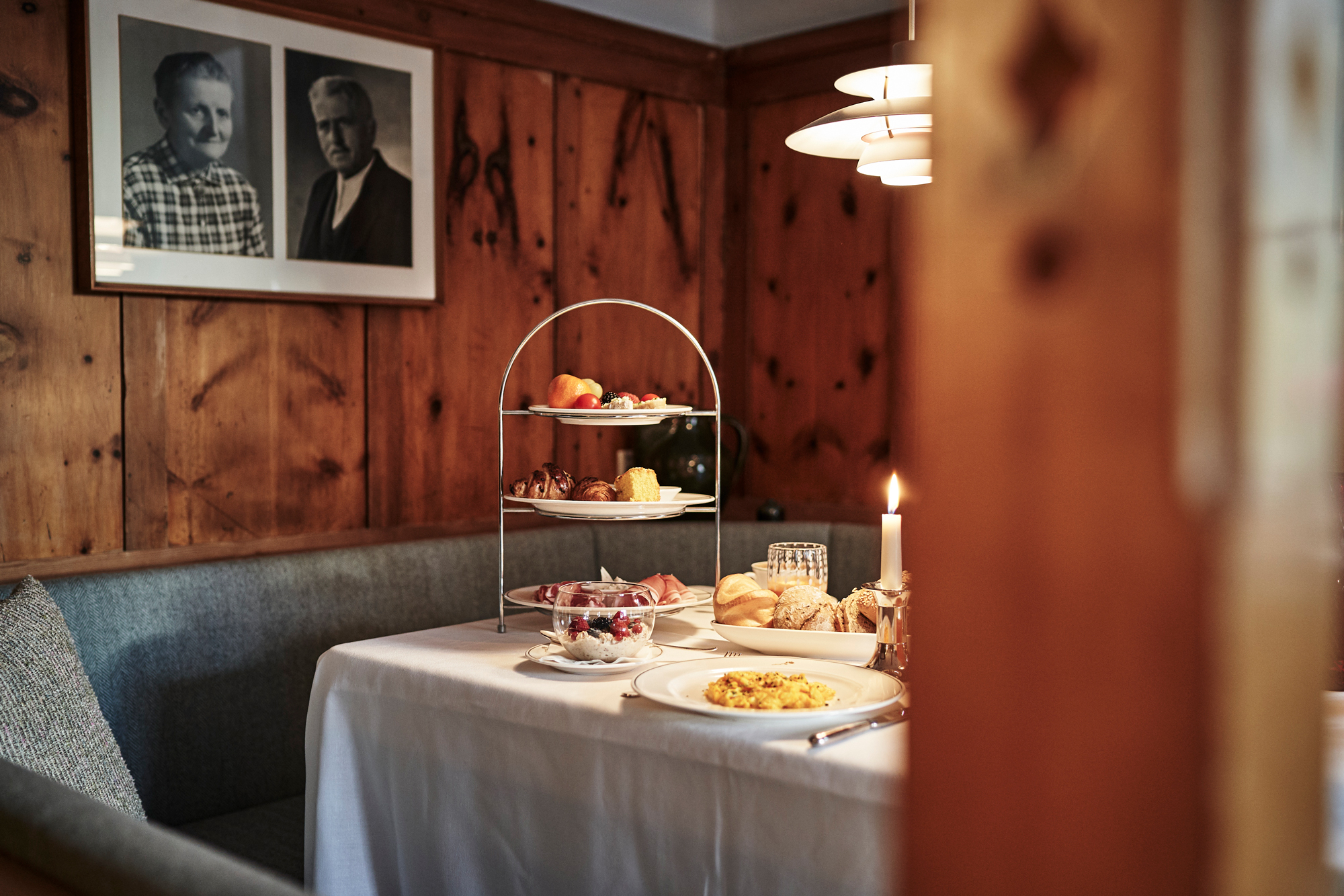 Table set for a meal with a tiered stand of pastries and fruits, candlelight, and vintage portraits on the wall.