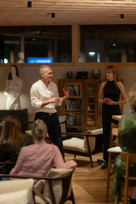 A man in a white shirt speaks while a woman in a black top stands beside him, addressing an audience in a cozy indoor sett...