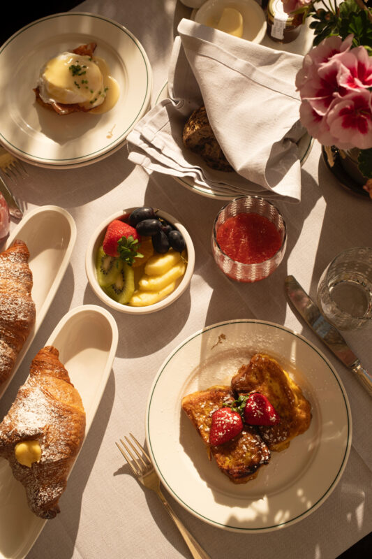 Brunch spread featuring croissants, French toast with strawberries, fruit, and drinks on a table with flowers.