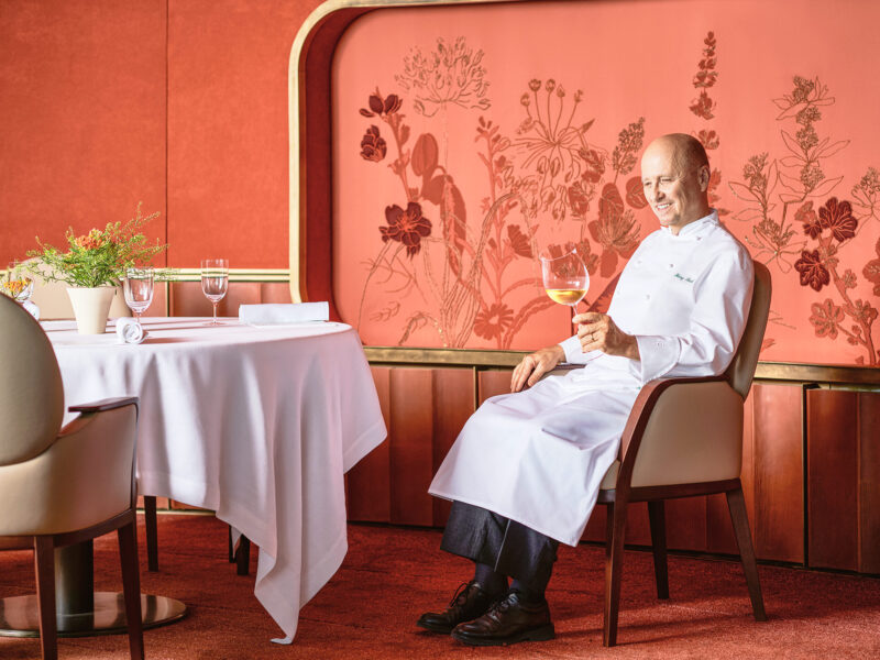Chef Heinz Beck seated in a dining room, holding a glass of wine, with floral decor on the wall behind him.