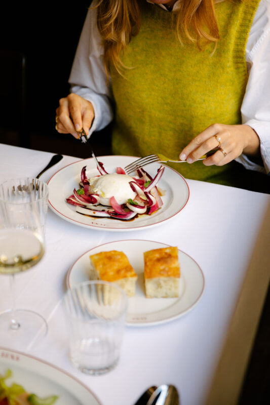 A person in a green sweater is enjoying a salad with poached egg, accompanied by bread, in a restaurant setting.