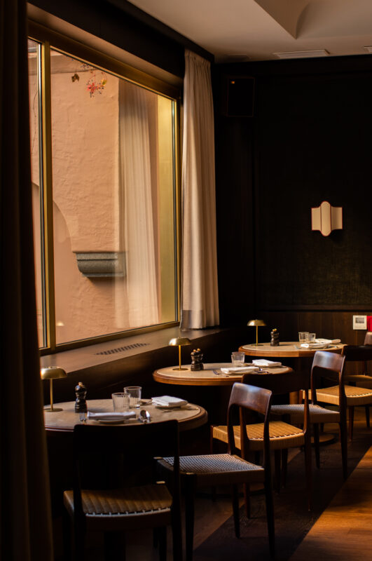 Elegant restaurant interior with wooden tables, chairs, and large windows letting in warm light.