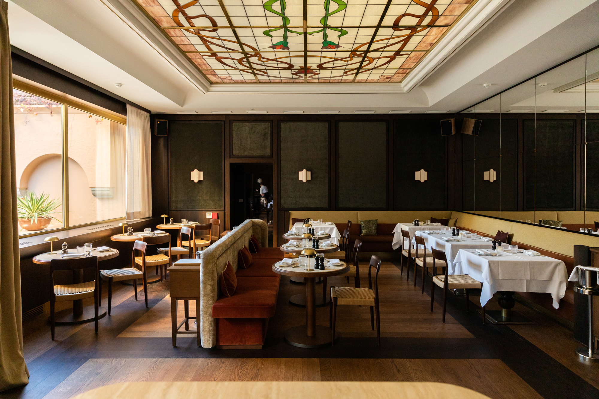 Elegant restaurant interior with wooden floors, tables set with white linens, and a stained glass ceiling.