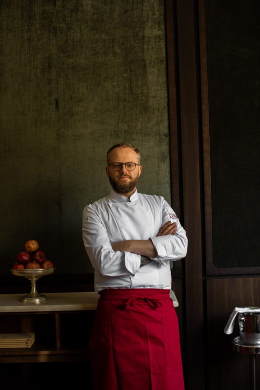 Chef in a white uniform and red apron stands confidently with arms crossed, against a textured green wall, apples on a tab...