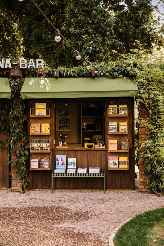 Wooden kiosk with shelves displaying brochures and magazines, surrounded by greenery at Parkhotel Mondschein.