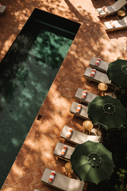 Lounge chairs with green umbrellas surround a pool, casting shadows on a brick patio at Parkhotel Mondschein.