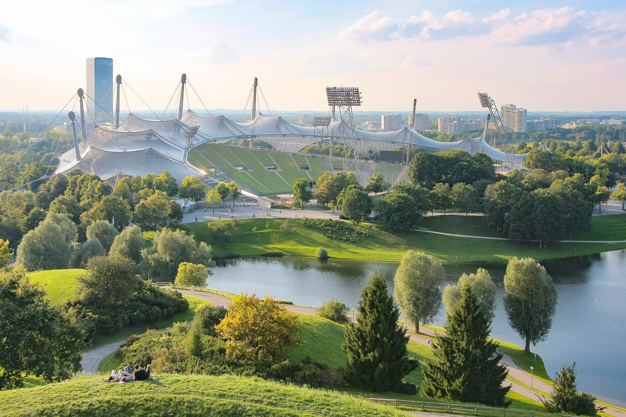 Olympiastadion in Munich, surrounded by greenery and a lake, with a clear sky and distant city skyline.