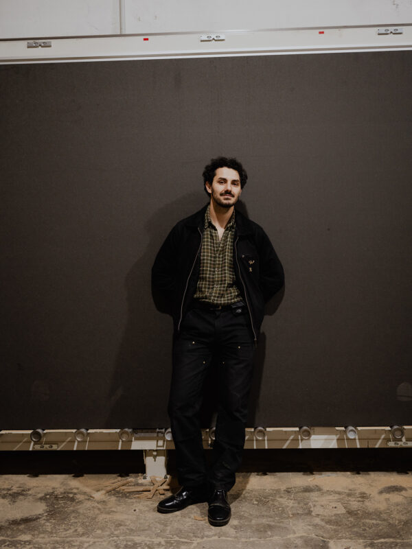Marco Zebrowski stands casually against a dark backdrop, wearing a checkered shirt and black pants, in an industrial setting.