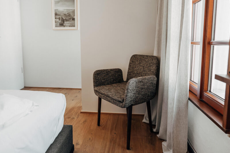 Cozy corner featuring a textured armchair next to a bed and a window with light curtains in a room.