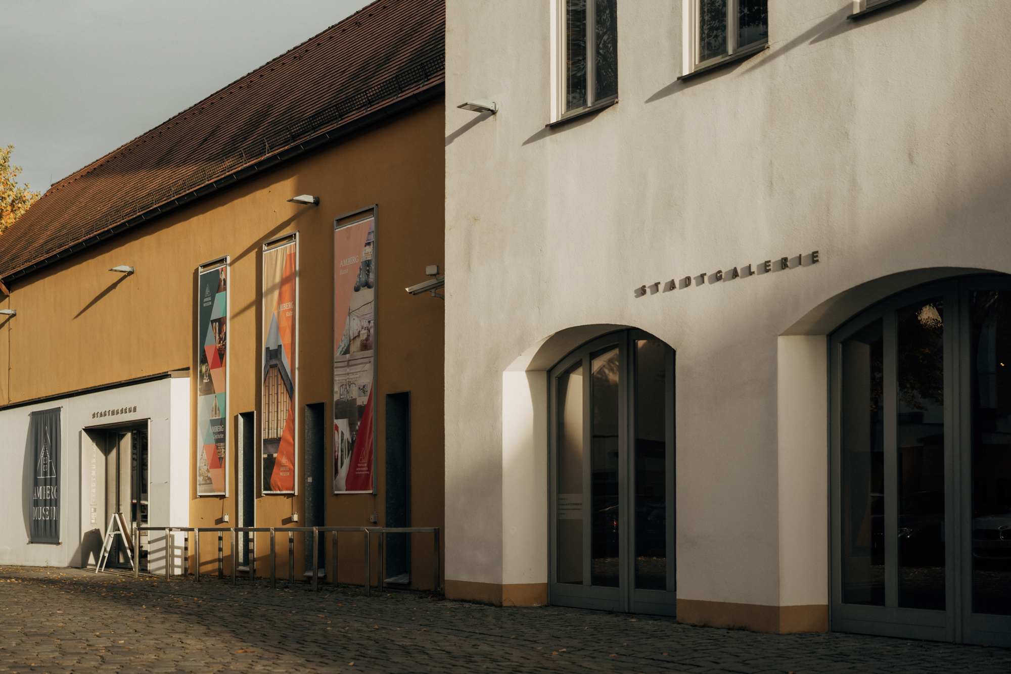 Two adjacent buildings with contrasting colors, one orange and one white, featuring large posters and a sign reading "Stad...