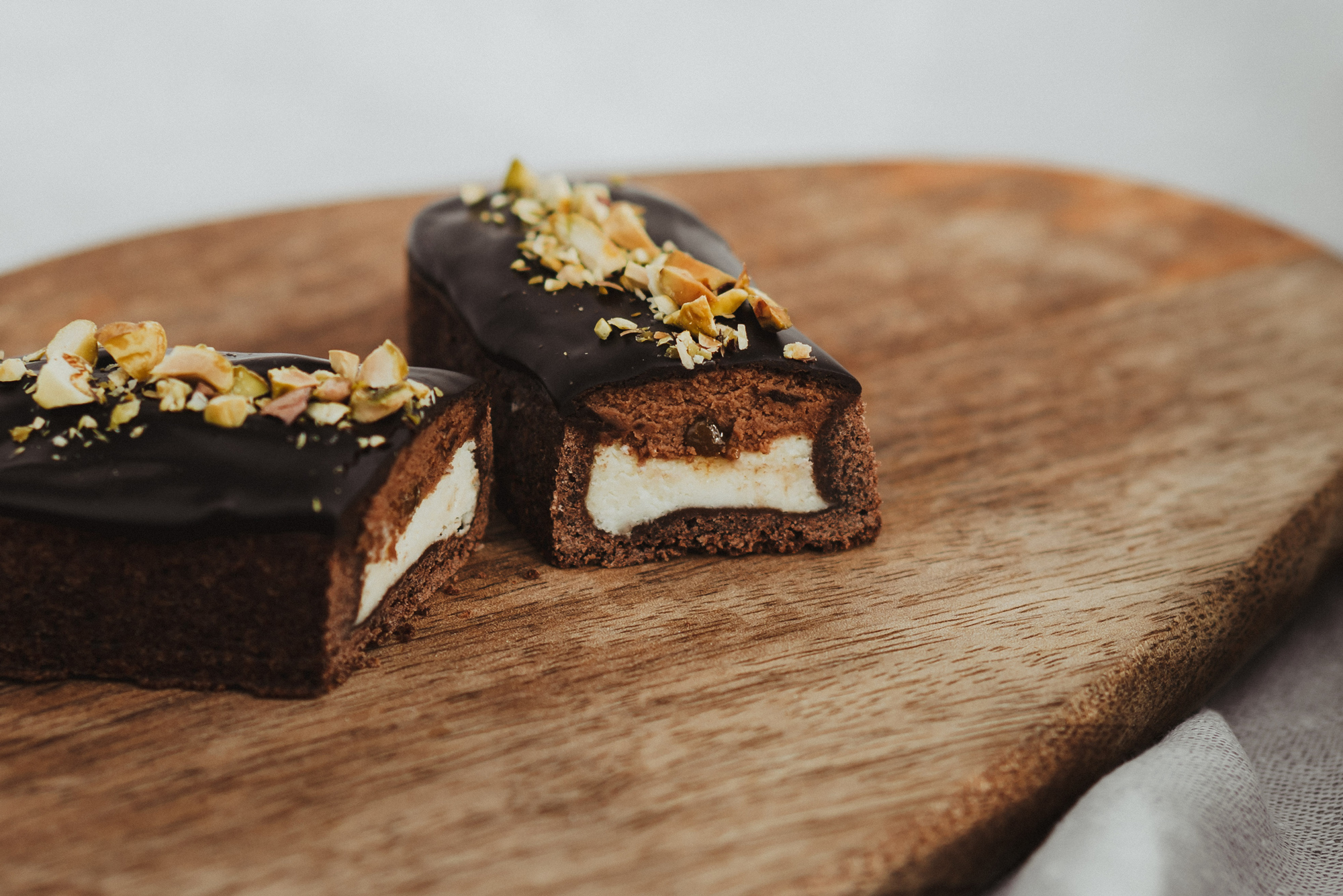 Chocolate-covered dessert bars with a creamy filling, topped with chopped pistachios, on a wooden serving board.