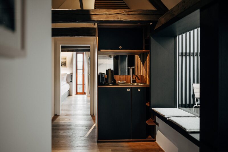 Modern interior of a cozy space featuring a kitchenette with dark cabinetry, wooden accents, and a view into a bedroom area.