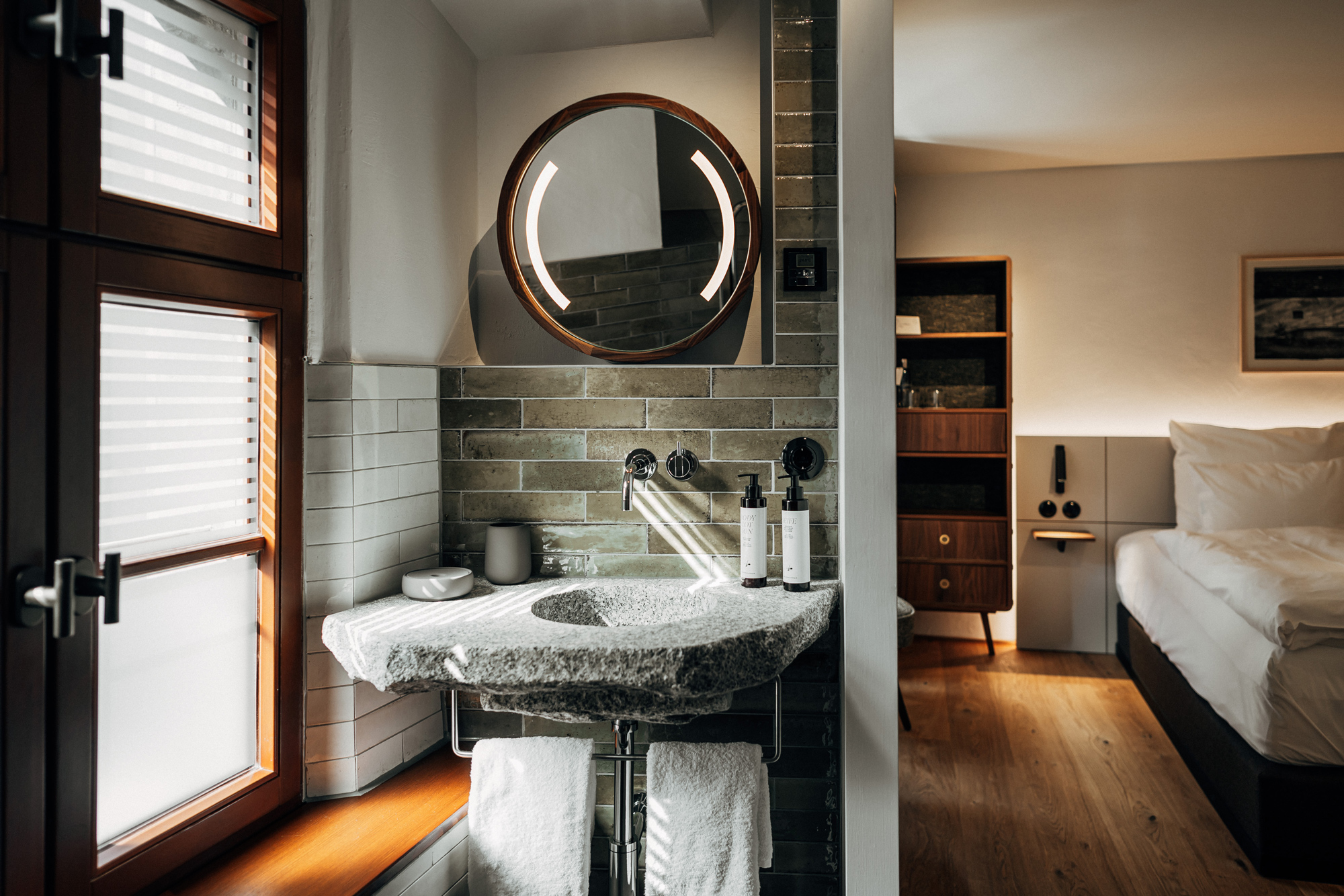 Modern bathroom featuring a stone sink, circular mirror, and toiletries, with a cozy bedroom visible in the background.