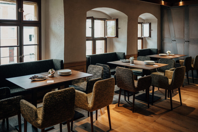 Cozy restaurant interior with wooden tables, upholstered chairs, and large windows allowing natural light.