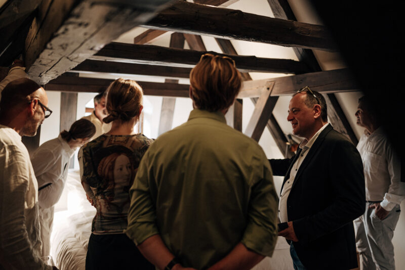 Group of people in a rustic attic space, engaged in conversation, with wooden beams visible in the background.