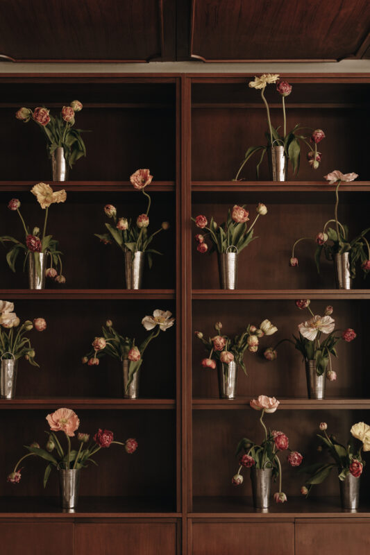 Vases with various flowers arranged on wooden shelves in a warm-toned interior setting.