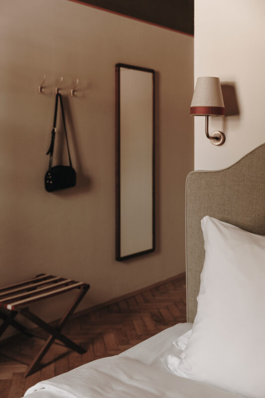 Cozy hotel room with a bed, a mirror on the wall, a wall lamp, and a bag hanging on hooks.