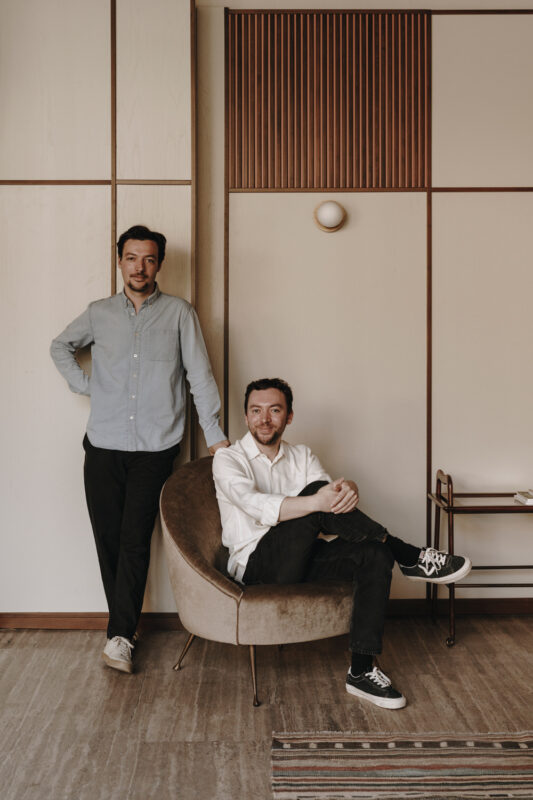 Two men pose in a stylish hotel room, one standing beside a modern chair while the other sits casually.