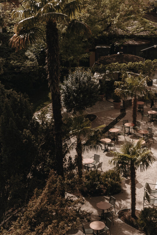 Lush garden area at Parkhotel Mondschein with palm trees and outdoor tables arranged on a stone patio.