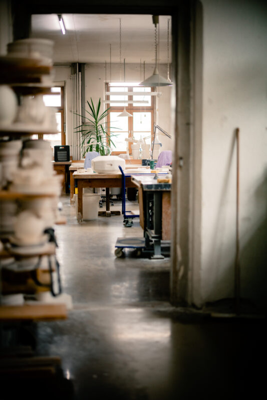 Artistic pottery studio with worktables, tools, and a large plant, viewed through a doorway with shelves in the foreground.