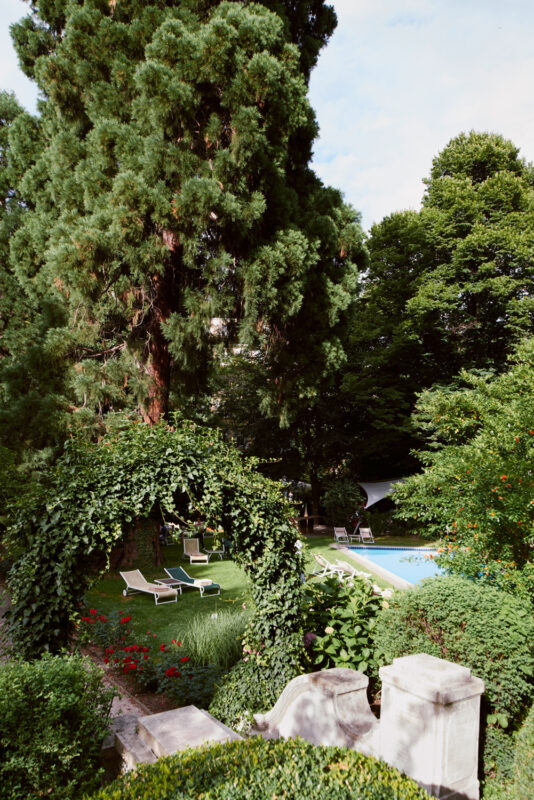 Lush garden with a pool, surrounded by tall trees and lounge chairs, framed by a green archway.