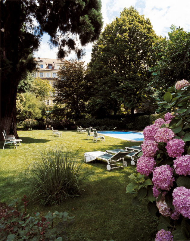 Lush garden at Parkhotel Laurin featuring a swimming pool, lounge chairs, and blooming hydrangeas.