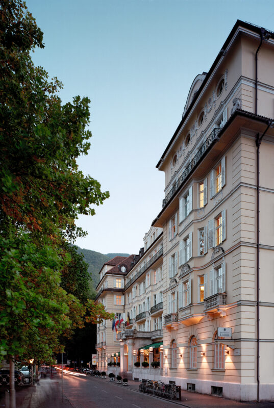 Elegant facade of Parkhotel Laurin, featuring balconies and decorative elements, framed by trees and a mountain backdrop.
