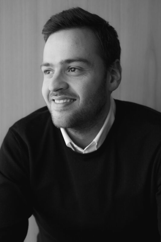 Smiling man in a black sweater, sitting against a wooden background, looking thoughtfully to the side.