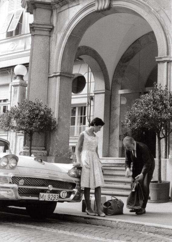 A woman in a white dress stands beside a vintage car while a man kneels, adjusting his bag, near the Parkhotel Laurin entr...