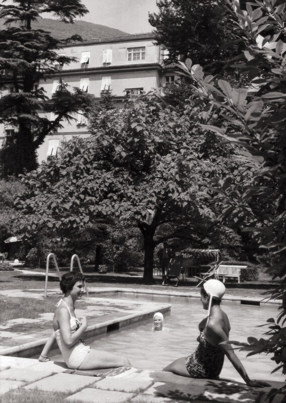 Two women in vintage swimsuits relax by a pool surrounded by trees at Parkhotel Laurin.