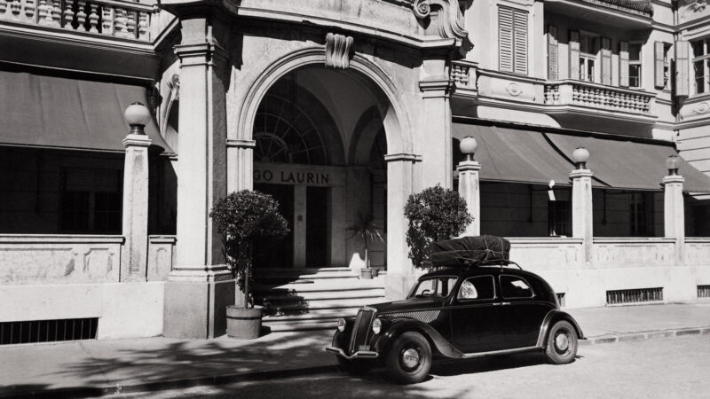 Classic black and white image of a vintage car parked outside the entrance of Parkhotel Laurin, featuring elegant architec...