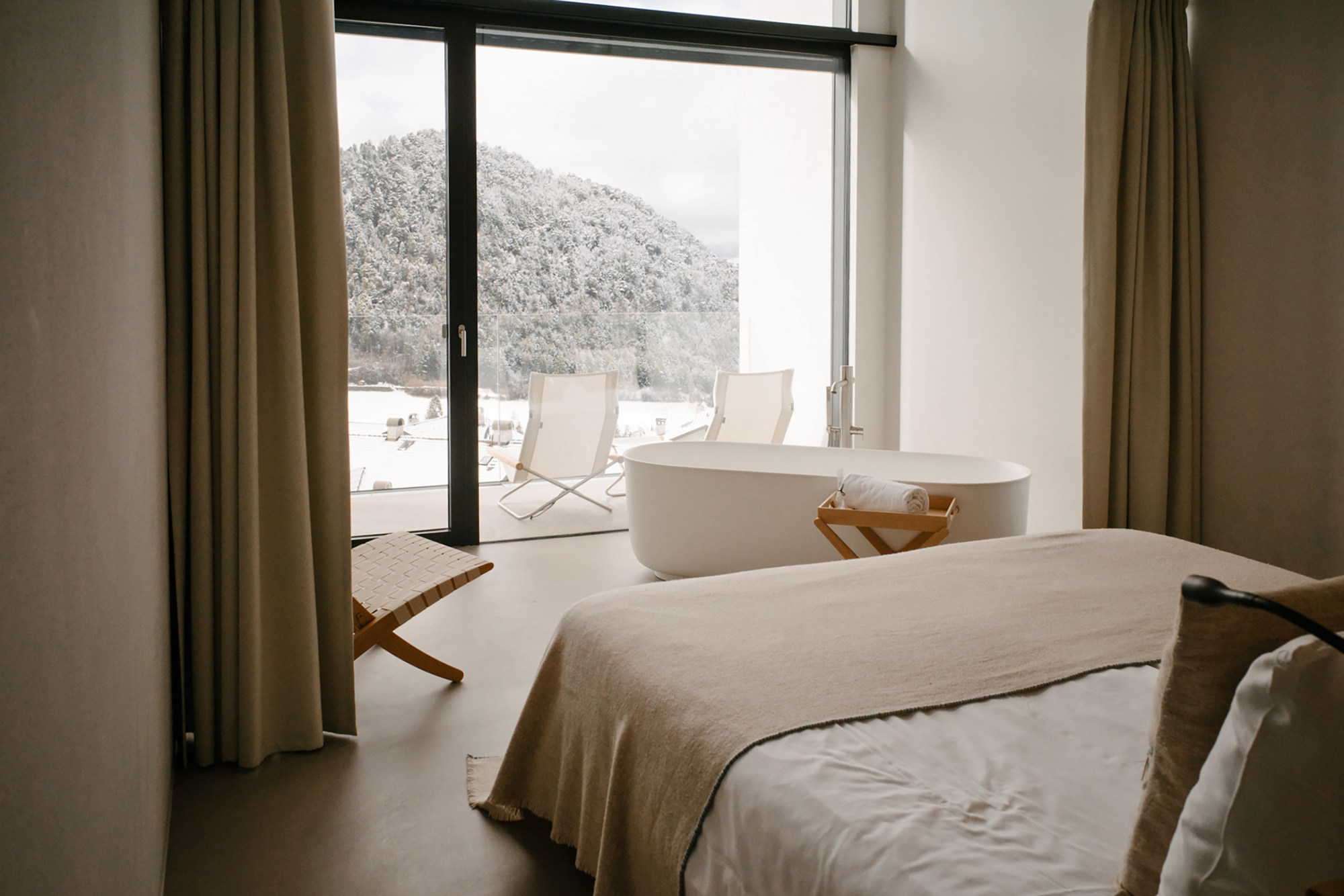 Modern hotel room with a bed, a freestanding bathtub, and large windows overlooking a snowy landscape.