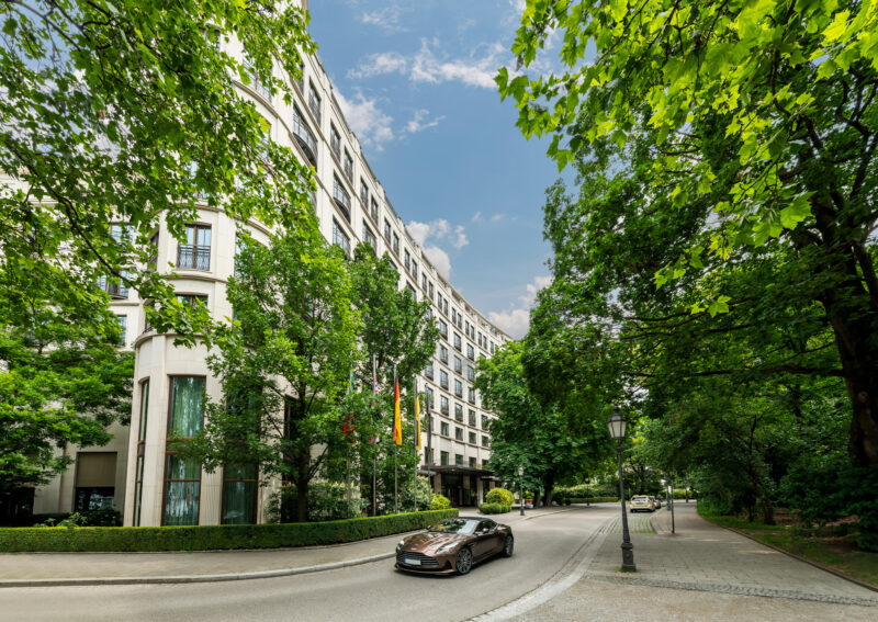 Modern hotel facade surrounded by lush greenery and trees, with a car driving along the curved road.
