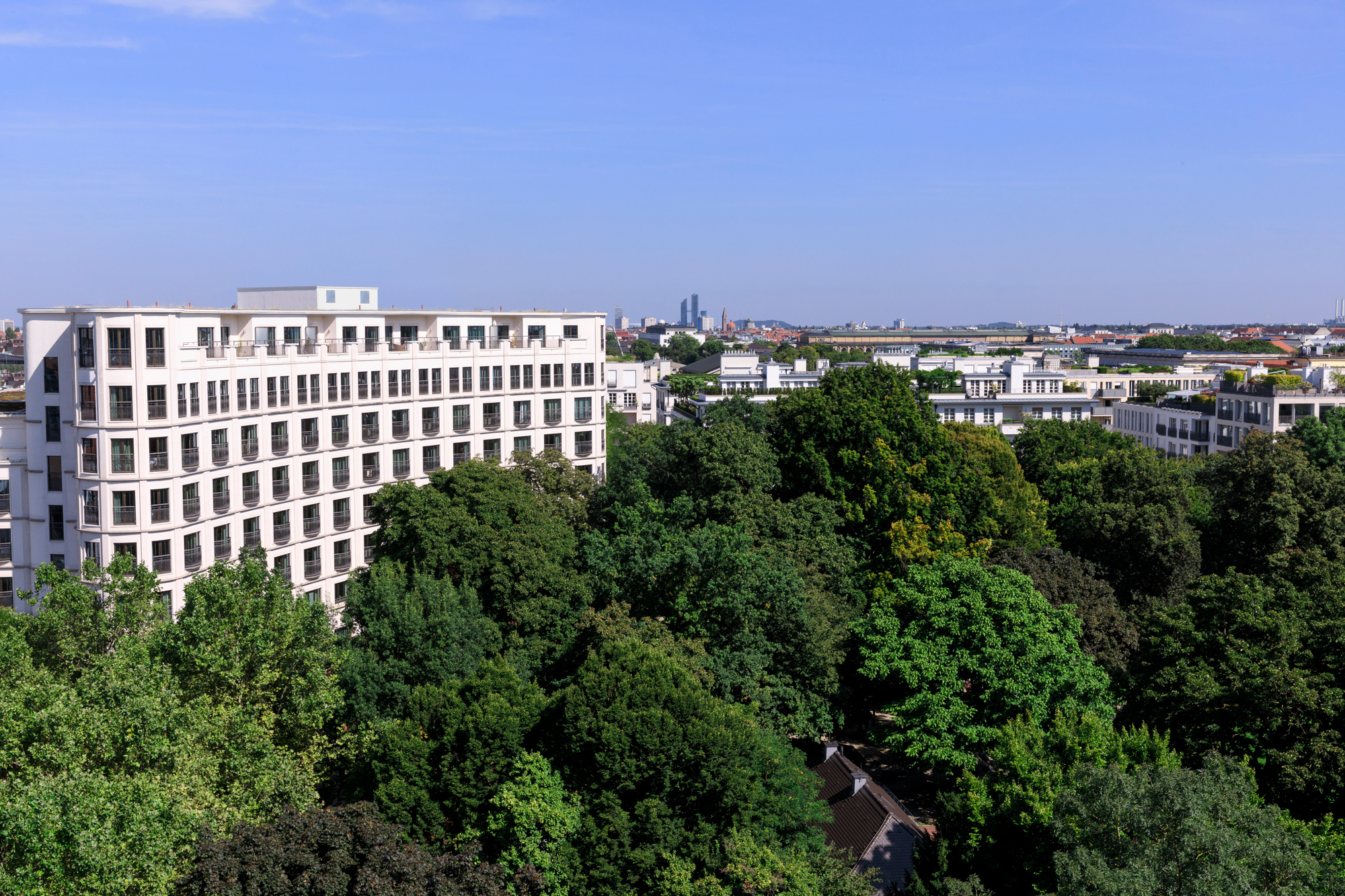 Modern hotel facade surrounded by lush greenery, with a clear blue sky and city skyline in the background.