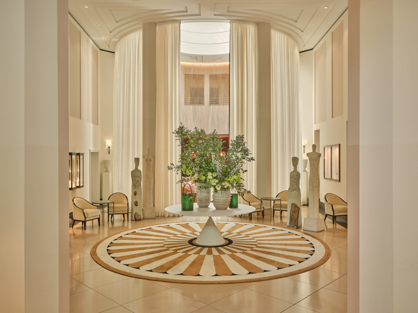 Elegant hotel lobby featuring a circular patterned floor, tall curtains, a central plant arrangement, and decorative sculp...