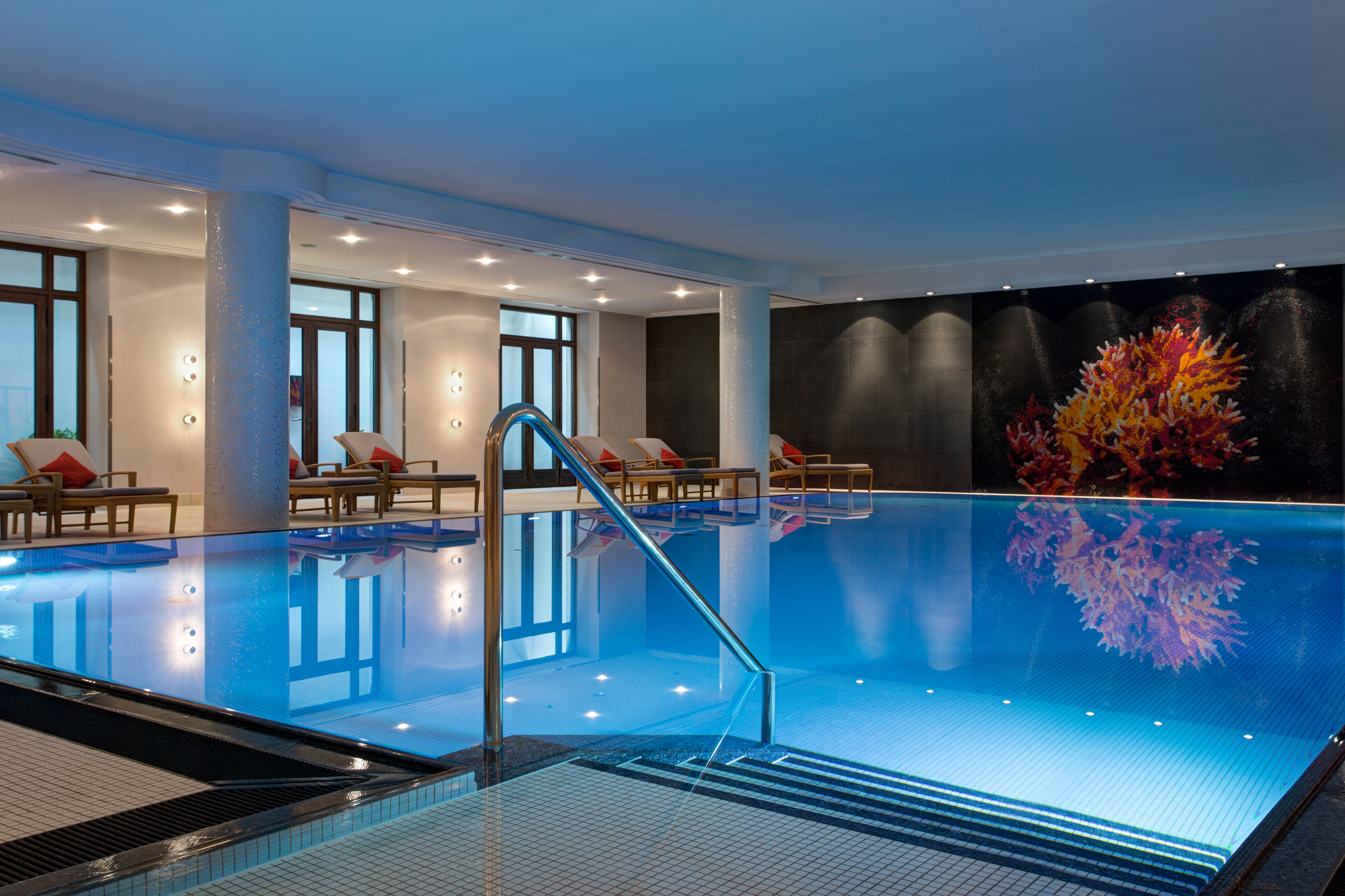 Indoor pool at The Charles Spa, featuring blue water, lounge chairs, and a coral mural on the wall.