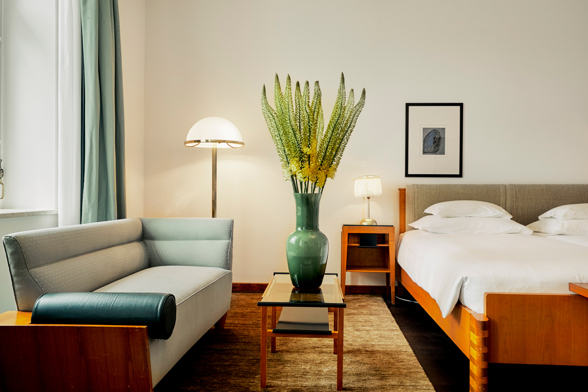 Modern hotel room featuring a bed, a green vase with flowers, a sofa, and a lamp.