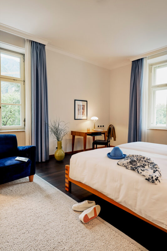 Modern hotel room with a double bed, a blue armchair, a desk, and a vase. Casual clothing and shoes are on the floor.