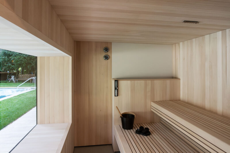 Wooden sauna interior with benches, a bucket, and a window overlooking a green lawn and pool area.