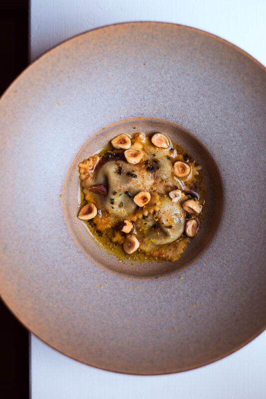 Ravioli dish topped with hazelnuts in a shallow, round beige plate, placed on a white surface.
